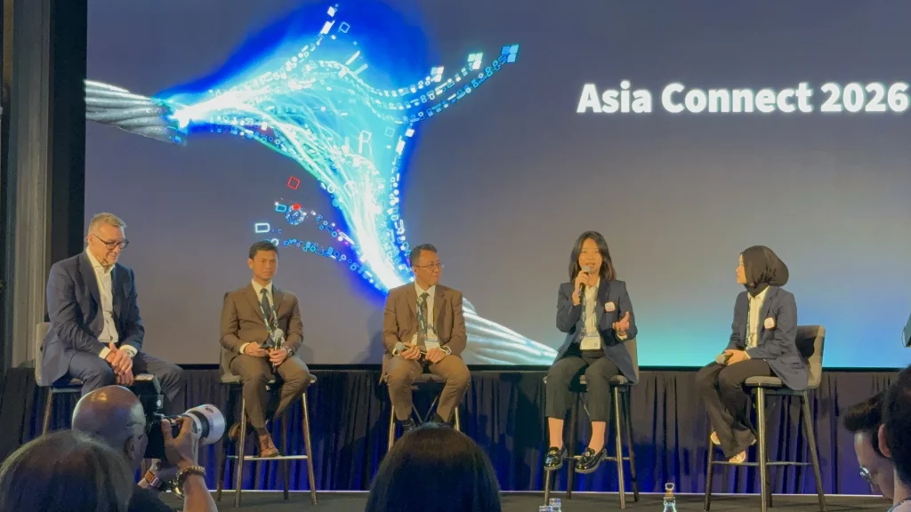 Lima narasumber profesional duduk di atas panggung dalam sesi diskusi panel bertema Asia Connect 2026. Seorang wanita di tengah sedang berbicara menggunakan mikrofon, berlatar belakang layar LED besar bertuliskan Asia Connect 2026. Sesi ini membahas transformasi digital industri manufaktur Indonesia bersama Epicor dan Prismatech